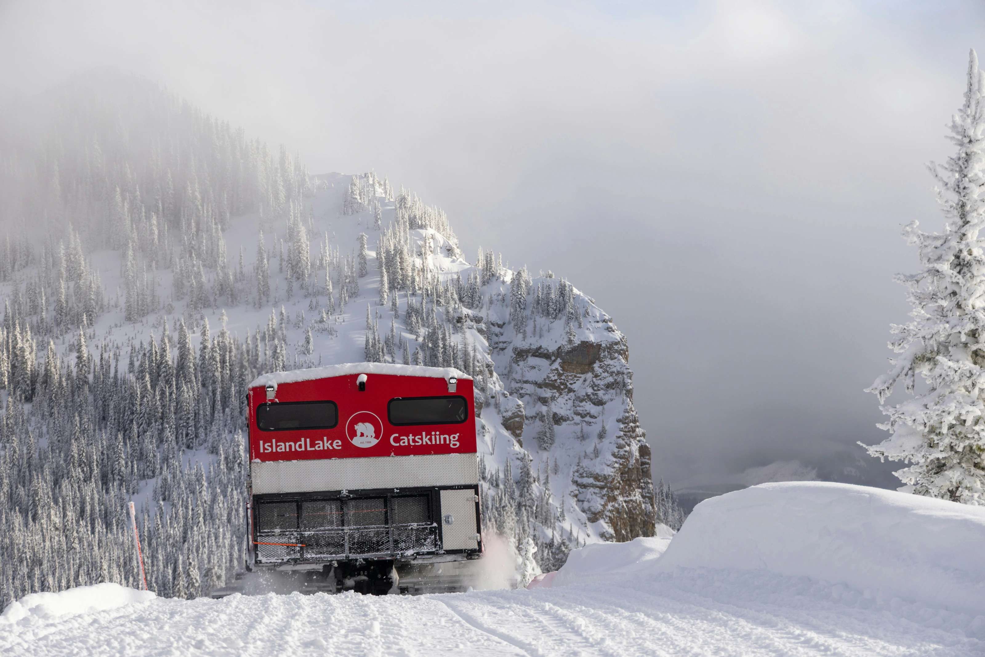 An Island Lake snowcat beneath a cliff lined with snow-covered trees