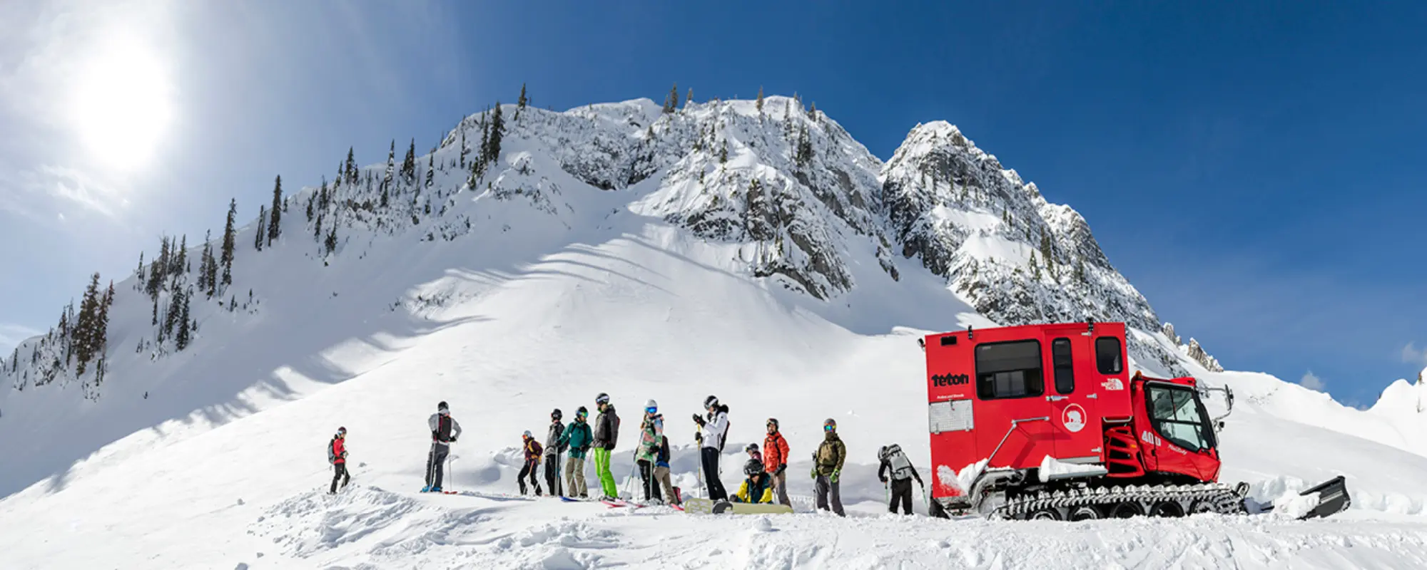 A group preparing to ski beside a red snowcat below a broad snow-covered mountain